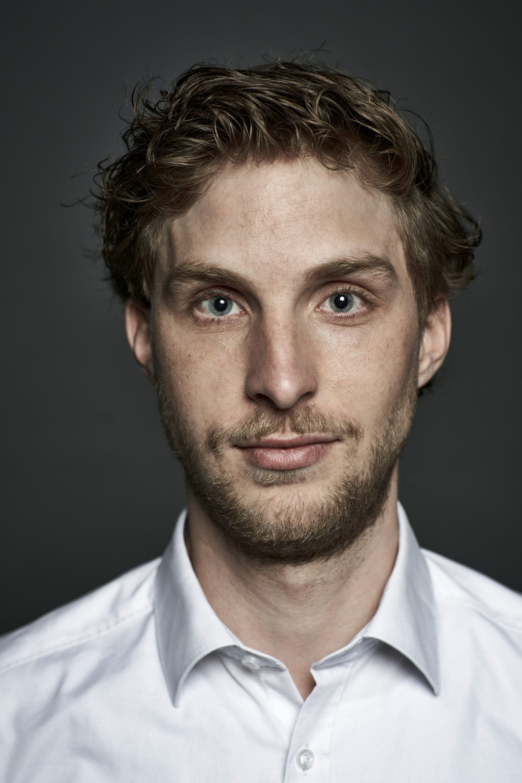 Christian Rammensee-Kink mit gewelltem hellbraunem Haar, blauen Augen und einem kurzen Bart trägt ein weißes Hemd und schaut mit neutralem Blick direkt in die Kamera.