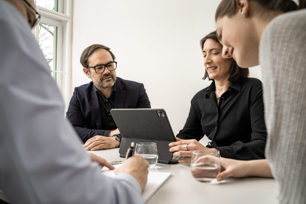 Philipp Wünsch, Sandra Markl und Laura Schneider sitzen in einem hellen Raum um einen Tisch herum und besprechen etwas, während sie auf ein Tablet schauen. Thomas Lehnert ist im Vordergrund verschwommen zu sehen und schreibt etwas mit einem Kugelschreiber auf ein Blatt Papier. Auf dem Tisch stehen zwei Gläser Wasser. Sie alle führen miteinander eine Besprechung.