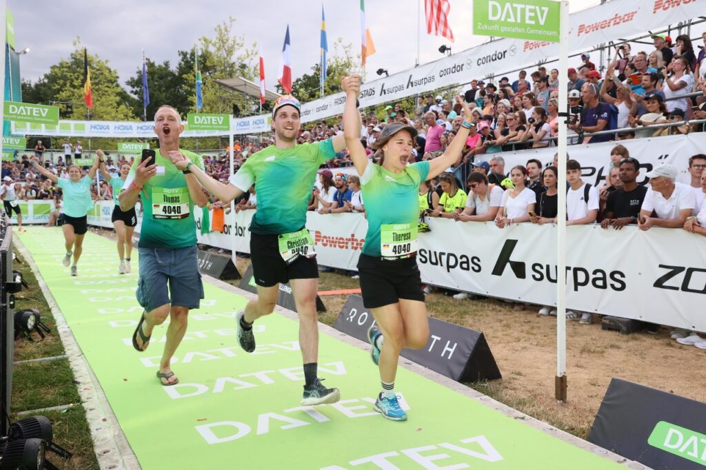 Drei Läufer von WLW bei der DATEV Challenge in Roth 2025 in passenden grünen Hemden, die sich an den Händen halten und lächeln, feiern, als sie die Ziellinie bei einem Rennen überqueren, mit jubelnden Zuschauern und Fahnen im Hintergrund.