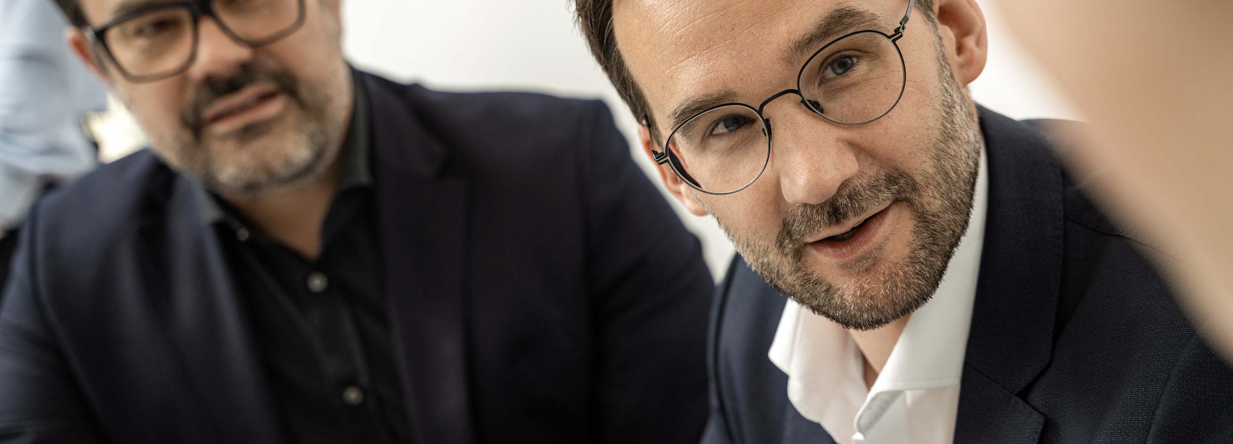 Philipp Wünsch und Andreas Selmer (v.l.) in Anzügen sitzen eng beieinander, hören aufmerksam zu und führen ein Gespräch mit einer dritten, nicht sichtbaren Person, die etwas verschwommen im Vordergrund zu sehen ist. Sie alle führen miteinander eine Besprechung.