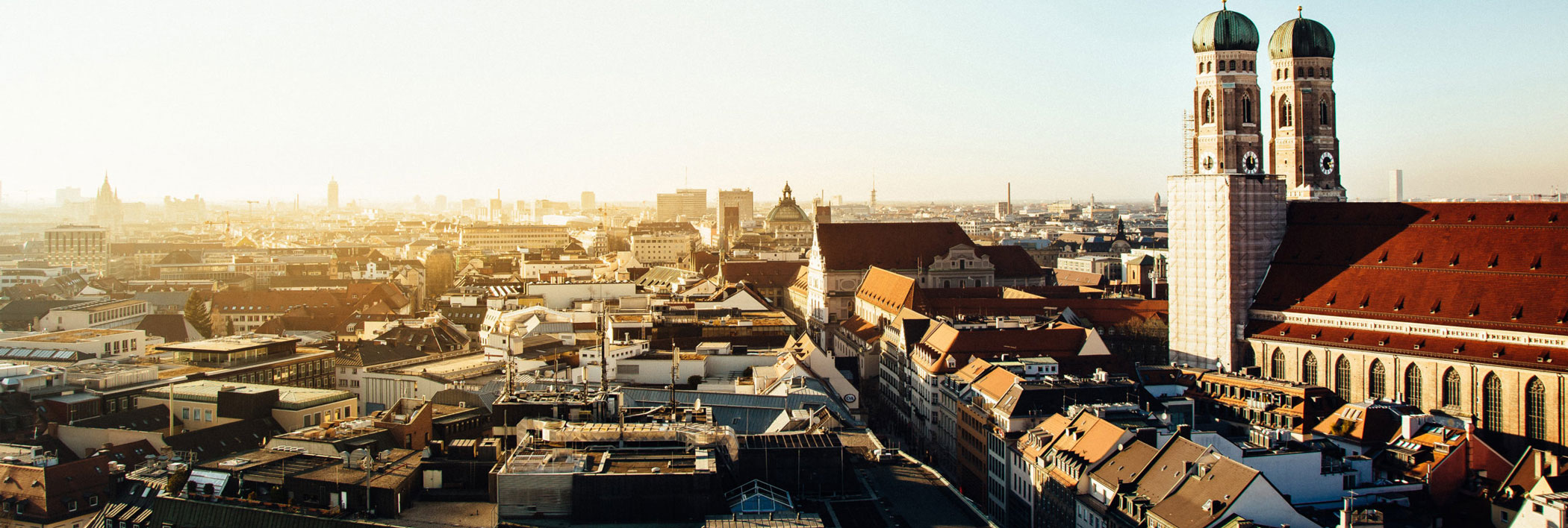 Ein Panoramablick auf München bei Sonnenuntergang, mit historischen Gebäuden und den Zwillingskuppeln der Frauenkirche, die sich über den Dächern der Stadt erheben.